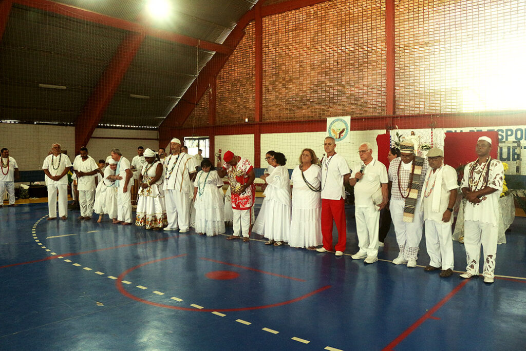 Lideranças da Associação Sábios da Paz reunidas em fila - uma pessoa ao lado da outra. Estão de branco, na Quadra de Esportes.